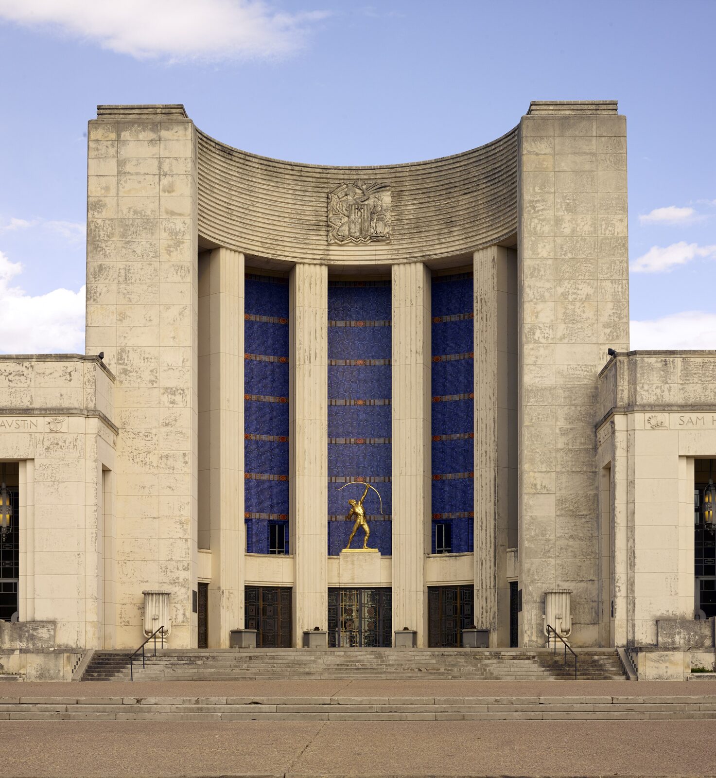 Limestone facade of the Hall of State at Fair Park in Dallas, with fluted columns framing a blue tile wall and a gold statue above the entrance