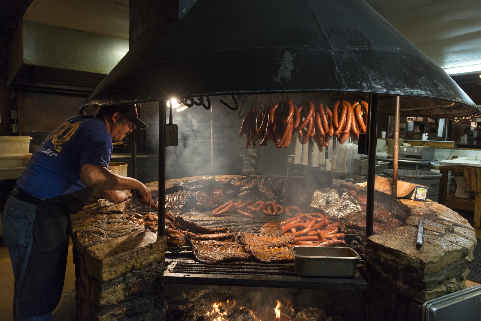 A pitmaster in a cap and apron tends ribs and sausage links on a large round stone barbecue pit with a wood fire burning below the grate