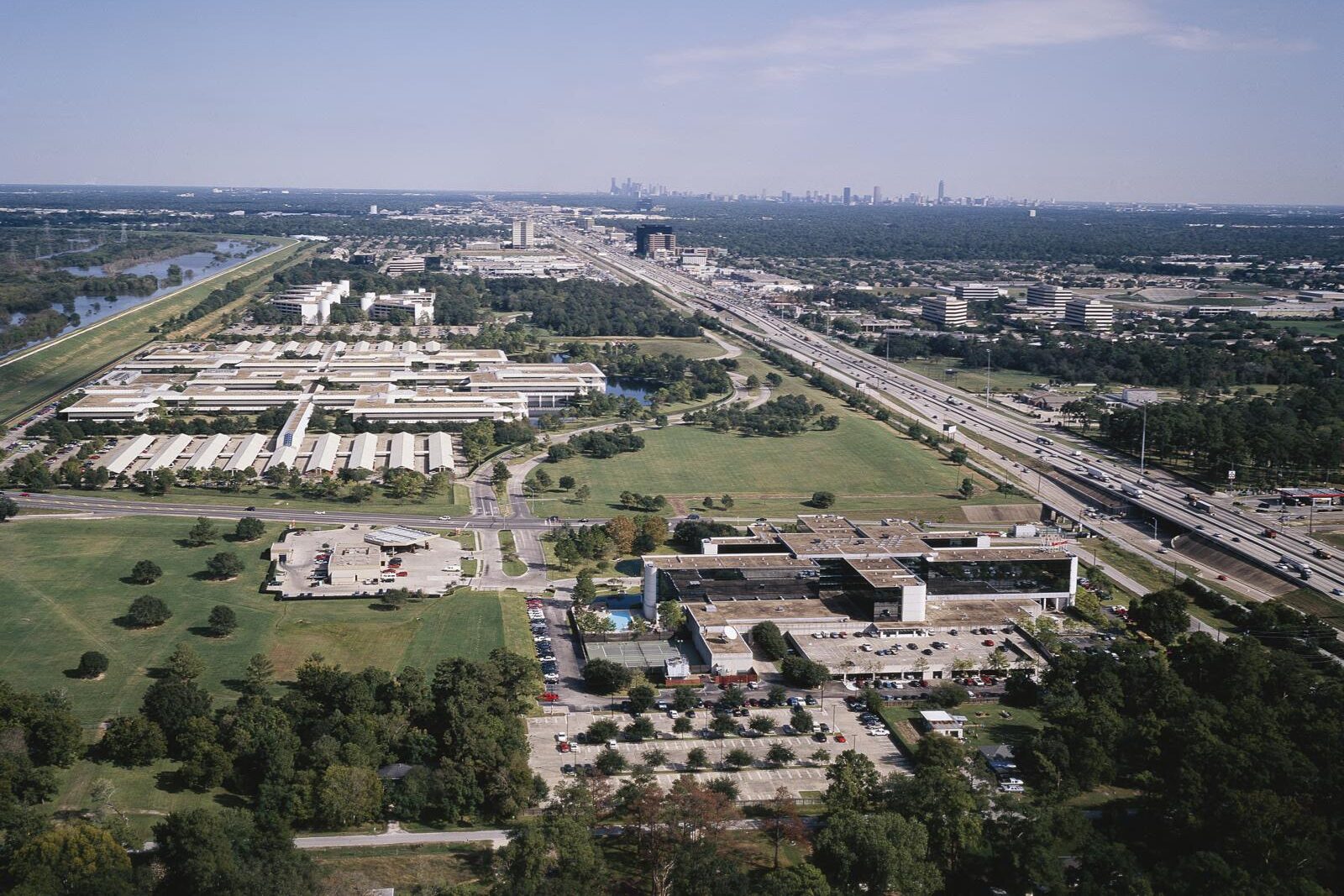 An aerial view of a wide multi-lane freeway running past office campuses toward a distant city skyline