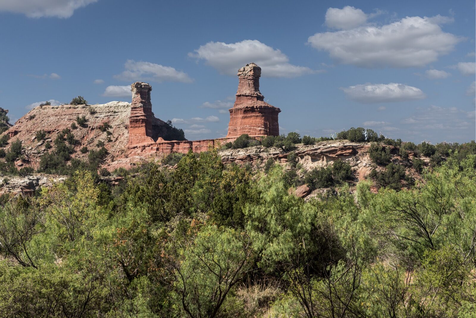 Two tall red sandstone rock formations rise above a ridge of layered rock and green brush under a blue sky in Palo Duro Canyon